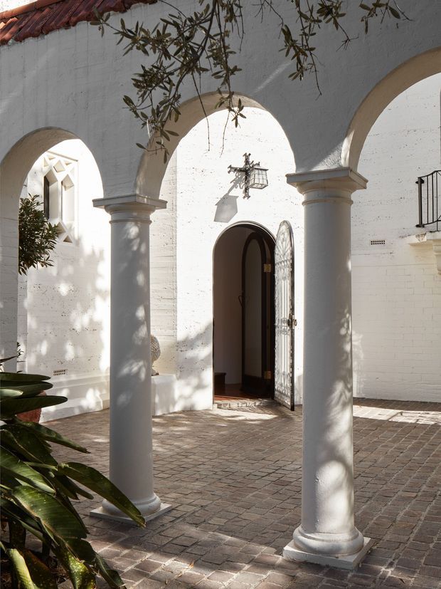 A sunlit Mediterranean-style courtyard or entry arcade with white stucco and brick walls, rounded arches supported by classical columns, a terracotta tiled roof edge, patterned brick paving, and a dark arched doorway with an ornate metal security gate. Olive branches cast shadows across the facade, while traditional lanterns and decorative details reinforce the historic, villa-like character.