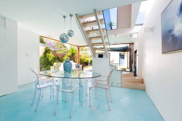 Bright contemporary open-plan dining area with a round glass dining table and transparent acrylic chairs, opening to a leafy courtyard through large glazed doors. The room features a vivid turquoise floor, white walls, mirrored and glass surfaces, sculptural chrome pendant lights, and an open timber stair with glass balustrade leading to an upper level. A skylight and clerestory glazing bring in abundant natural light.