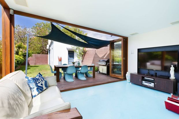 Bright contemporary living room opening directly onto a timber deck through wide timber-framed glazed folding or sliding doors. The room has a pale blue floor, white walls and ceiling, a white sofa with patterned cushions, and a dark timber media console with a wall-mounted television. Outside is a dining setting, barbecue, and sculptural shade sail, with a garden and neighbouring structures beyond.