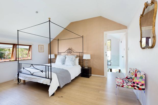 Bright bedroom with a black metal four-poster canopy bed, white bedding, light timber flooring, and a warm tan feature wall behind the bed. Large horizontal windows bring in natural light, while a colourful patterned accent chair and ornate gold-framed mirror add decorative contrast. An open doorway leads to an adjoining area, possibly a bathroom, but the bedroom is the primary space.