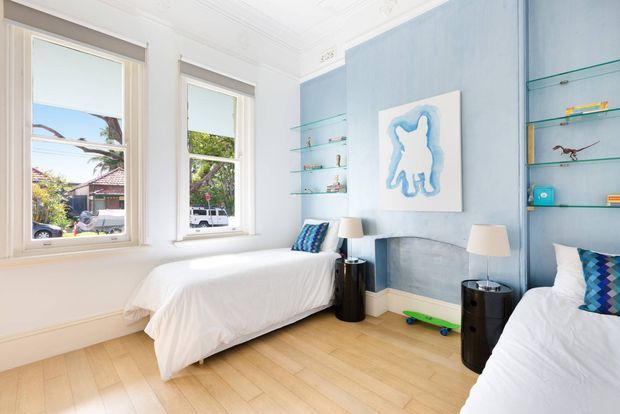 Bright twin bedroom with two single beds, pale timber flooring, white bedding, and a soft blue feature wall. Built-in glass display shelving, large traditional sash windows, bedside tables, and playful blue patterned cushions create a clean, airy shared children's or guest bedroom. A fireplace or chimney breast is integrated into the blue wall beneath artwork.