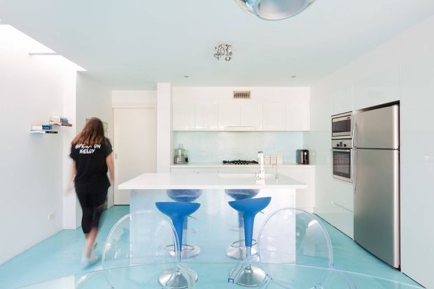 Bright, compact contemporary kitchen with glossy white cabinetry, a white island bench, pale aqua glass splashback and matching aqua flooring. The island has two vivid blue sculptural stools, while transparent acrylic dining chairs and a glass table occupy the foreground. Integrated double ovens and a stainless-steel refrigerator are visible on the right.