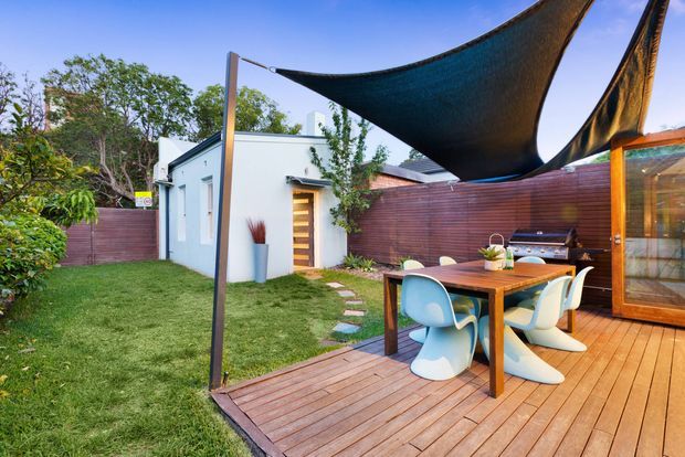 A landscaped backyard entertaining area with a timber deck, outdoor dining table and sculptural chairs beneath large triangular shade sails. The deck adjoins a timber-clad outdoor structure or glazed garden room, with a built-in barbecue against a horizontal timber privacy fence. A small white outbuilding, lawn, stepping stones and established planting complete the contemporary outdoor setting.