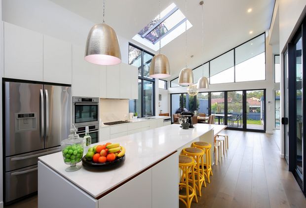 Bright, spacious contemporary kitchen with a large white island, integrated white cabinetry, stainless-steel refrigerator and oven, pale timber flooring, and multiple metallic pendant lights. Floor-to-ceiling glazing, a skylight, and clerestory windows bring in abundant natural light, with a dining or living area visible beyond the kitchen.