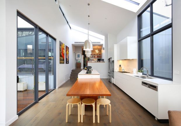 Bright contemporary open-plan kitchen viewed from the dining end, with a long timber dining table and stools leading to a central island. White handleless cabinetry, integrated appliances, a pale benchtop, large black-framed windows and doors, exposed pendant lighting, and multiple roof skylights create an airy, high-ceilinged space. A living or outdoor area is visible through the glazing.