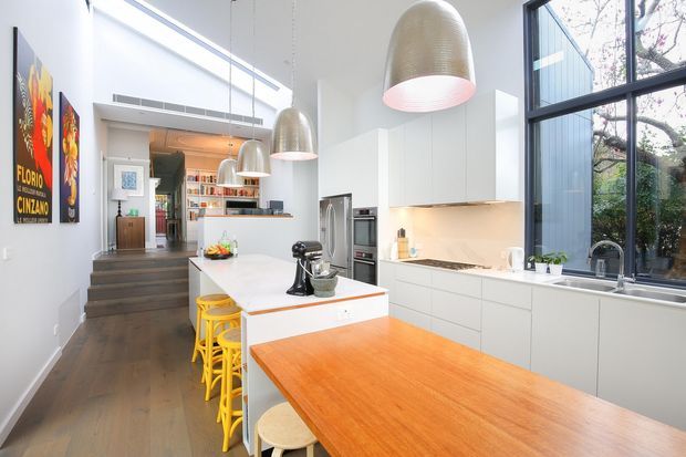 Bright, elongated contemporary kitchen with a large white island, yellow stools, integrated stainless-steel appliances, white cabinetry, and a warm timber dining bench extension. Oversized woven pendant lights hang above the island, while tall black-framed glazing brings in abundant natural light and views of trees. Dark timber flooring and colourful wall art add warmth and character.