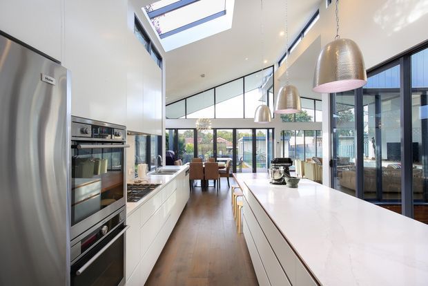 Bright, spacious contemporary kitchen with a long white island, extensive white built-in cabinetry, integrated stainless-steel appliances, timber flooring, and multiple metallic pendant lights. The kitchen opens to a dining and living area through large black-framed glazed partitions and doors. A dramatic high ceiling includes a large skylight, creating an airy open-plan interior.