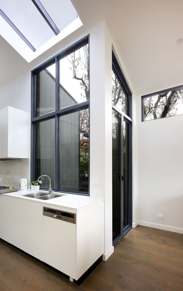Bright contemporary kitchen interior with a compact white island featuring an integrated double sink and dishwasher, tall dark-framed glazing, a skylight, white walls, and warm timber flooring. The high windows and rooflight bring in abundant natural light, with trees visible outside.