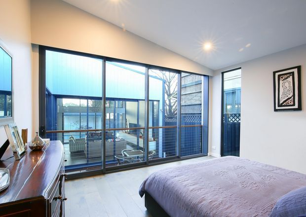 Bright contemporary bedroom with a large bed, pale textured coverlet, white walls and light flooring. A full-height glazed wall with dark frames overlooks an enclosed courtyard or terrace, while a timber dresser and framed artwork add warmth. Recessed ceiling lights and extensive glazing create an airy, modern interior.