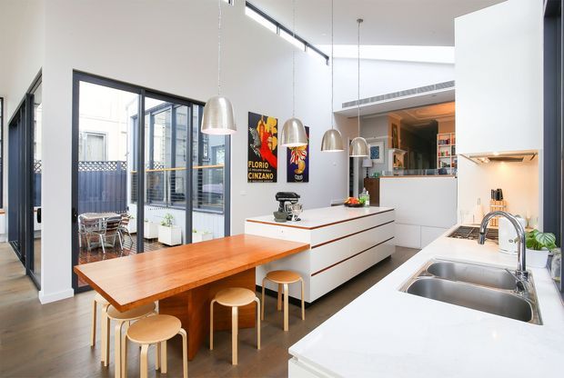 Bright open-plan kitchen with a long white island and integrated cabinetry, a timber dining extension with stools, stainless-steel double sink, white benchtops, pendant lights, and extensive black-framed glazing opening toward a small courtyard. The space has a clean contemporary fit-out with warm timber accents and artwork.