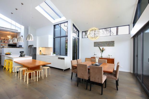 Bright, open-plan contemporary kitchen and dining area with a high ceiling, extensive dark-framed glazing, and a roof skylight. The kitchen features white fixed cabinetry, a timber island with casual yellow stools, pendant lights, and integrated appliances. A separate timber dining table with upholstered chairs sits beside a timber sideboard, while a sculptural chandelier and indoor planting add visual accents.