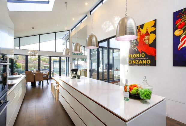 Bright, open-plan contemporary kitchen with a long white island, integrated cabinetry, timber flooring, large black-framed windows and clerestory glazing. The island includes seating and a sink, with sculptural pendant lights above. A dining area and garden-facing glazing are visible beyond the kitchen, along with colourful vintage-style wall artwork and countertop accessories.