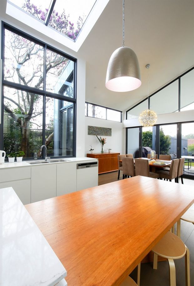 Bright open-plan kitchen and dining area with a large timber dining table in the foreground, white kitchen cabinetry and a pale stone benchtop, warm timber sideboard, black-framed floor-to-ceiling glazing, and a dramatic glazed skylight. Sculptural pendant lights hang above the kitchen and dining zones, with a leafy outdoor view visible through the windows.