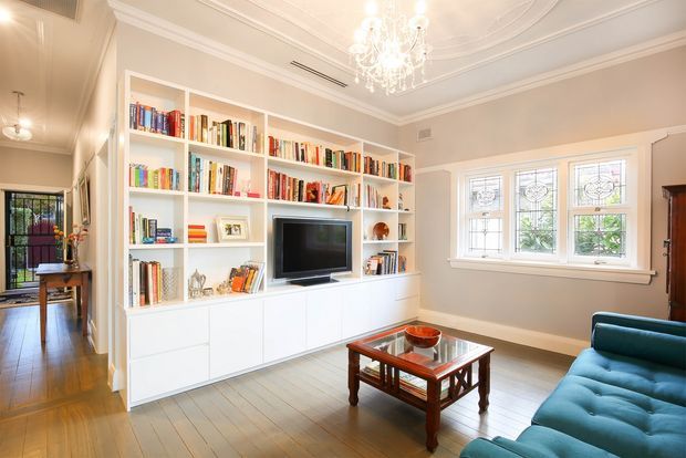 A refined living room with built-in white shelving and cabinetry spanning the main wall, filled with books and centred around a television. The room features a teal upholstered sofa, a dark timber and glass coffee table, pale timber flooring, decorative leaded-glass windows, high ornate plaster ceilings, crown moulding, and a crystal chandelier. A glazed doorway and adjoining circulation space are visible to the left.