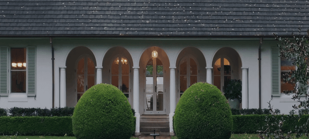 Symmetrical front facade of a refined traditional home, featuring a long dark shingle roof, white rendered walls, repeated arched openings supported by slender columns, tall timber-framed glazed doors, pale green shutters, and formal clipped topiary and hedge landscaping. The centered entrance and balanced composition create a stately, polished appearance.
