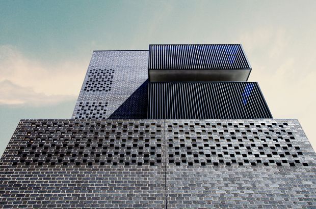 Upward-looking exterior architectural photograph of a tall contemporary building. The facade combines dark brick masonry with extensive patterned perforations and projecting upper volumes clad in dark vertically ribbed metal or glass. Strong geometric composition, sharp shadow lines, and a pale sky create a dramatic urban presentation.