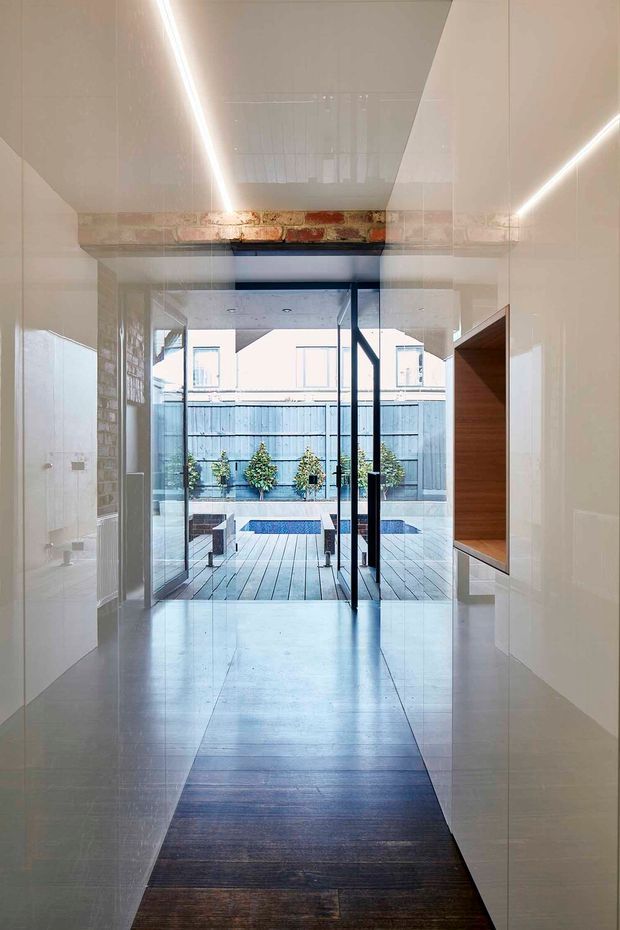 A narrow, minimalist interior corridor leading through full-height glazed doors to a compact courtyard and swimming pool. The corridor has glossy white wall and ceiling surfaces, integrated linear LED lighting, a dark timber floor runner, exposed brick detailing at the ceiling transition, and a recessed timber-lined wall niche on the right. The view opens to a paved outdoor area with a small pool and planted boundary wall.