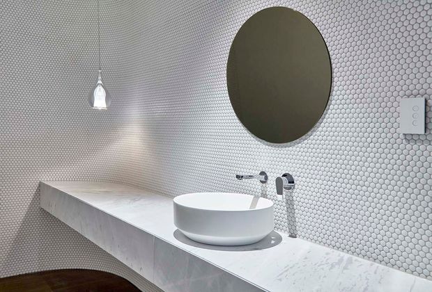 A refined contemporary bathroom vanity with a floating white marble or stone benchtop, vessel basin, wall-mounted chrome tapware, and a large round mirror. The walls are clad in small circular white and grey mosaic tiles, with a sculptural pendant light and minimalist switch plate adding detail.