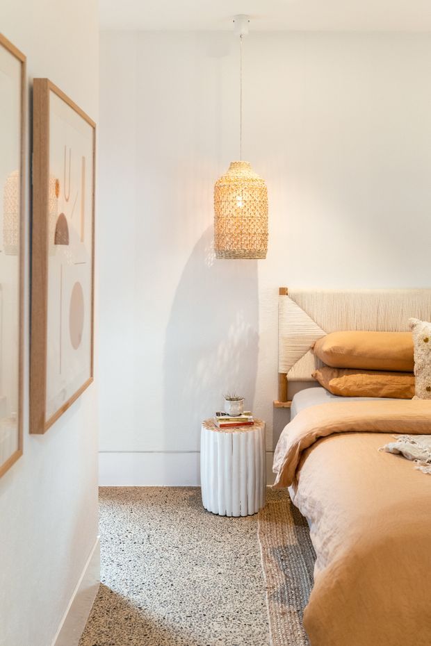 A softly lit, warm-toned bedroom corner featuring a textured upholstered bed with tan bedding and pillows, a woven pendant light, terrazzo flooring, framed abstract artwork, and a small fluted white side table. The palette is calm and natural with cream, beige, tan, and light timber accents.