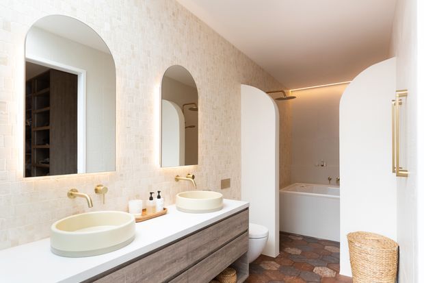 Bright, warm-toned bathroom with a long double vanity, two round vessel basins, brushed gold wall-mounted taps, illuminated arched mirrors, textured pale wall tiles, and a timber-fronted vanity. An arched opening leads to a separate bath and shower area with a freestanding-style bathtub and dark irregular stone-look floor tiles.