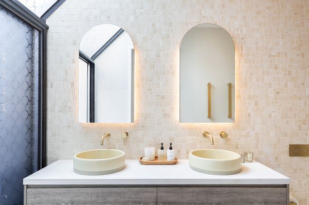 Contemporary double-basin bathroom vanity with two round vessel sinks, arched mirrors, warm brass tapware, pale mosaic wall tiling, and grey wood-grain cabinetry. A dark patterned glass shower screen is visible at left.
