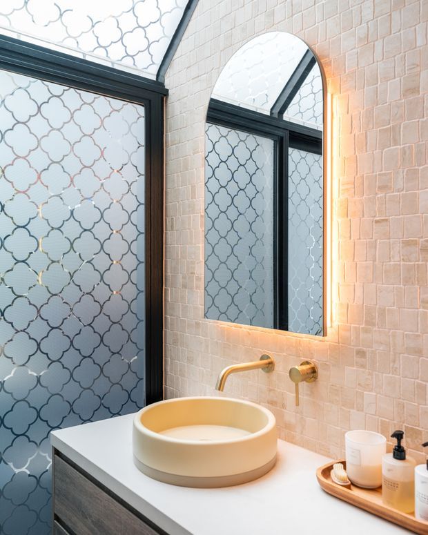 A contemporary bathroom vanity with a round vessel basin on a light benchtop, brushed brass wall-mounted tapware, a large arched mirror with warm backlighting, pale textured wall tiles, and black-framed patterned frosted glazing. The space has a refined, softly illuminated aesthetic with geometric detailing.
