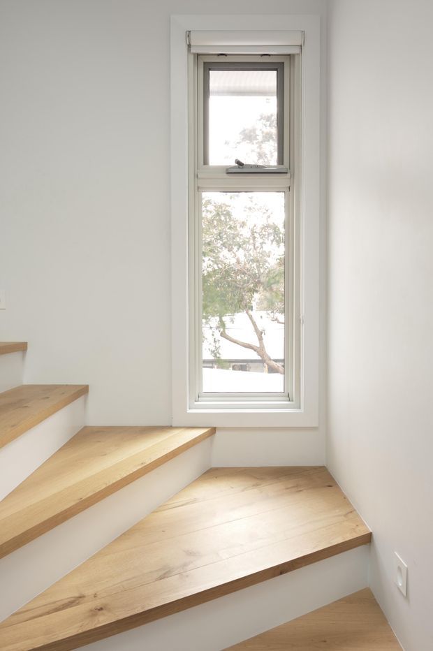 A bright, narrow stairway with pale timber treads and white risers, turning around a small landing. A tall white-framed window brings in natural light, with plain light walls and minimal detailing.