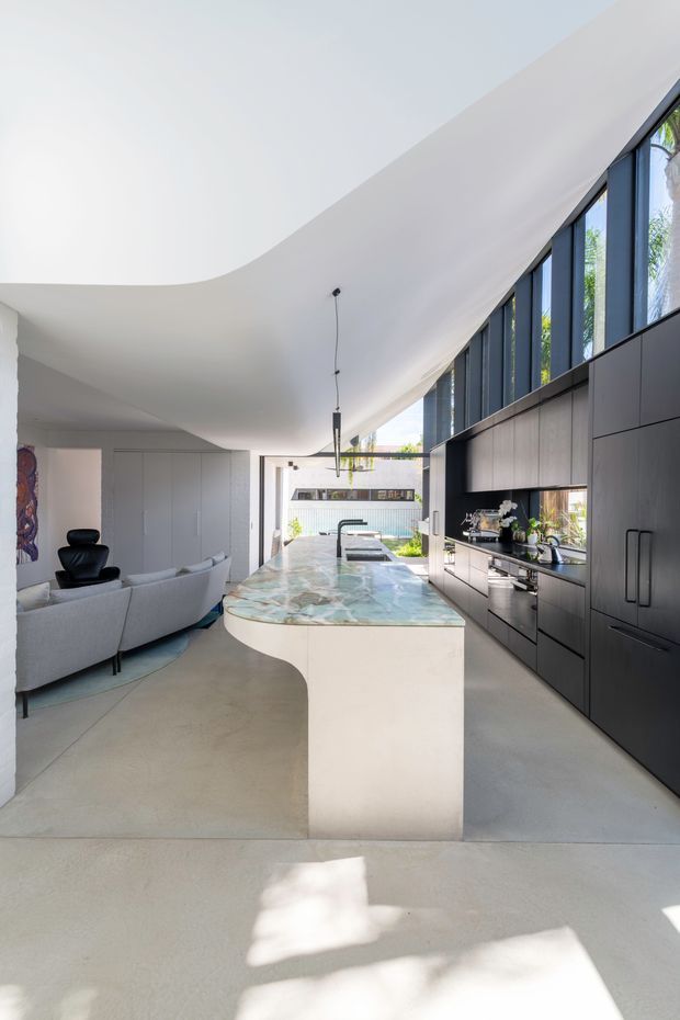 A bright, open-plan contemporary kitchen and living space. The kitchen features extensive matte dark cabinetry, integrated appliances, a large curved island with a pale veined stone benchtop, and a linear run of tall dark-framed windows. A sculptural curved white ceiling, polished concrete floor, and minimalist pendant lighting create an airy, modern interior connected visually to a living room and landscaped outdoor area.