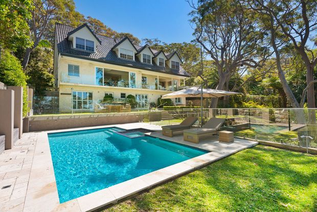 A large landscaped backyard with a rectangular swimming pool and integrated spa, surrounded by pale stone paving, glass pool fencing, sun loungers and umbrellas. Behind the pool is a substantial multi-storey residence with a pale rendered facade, steep dark tiled gable roof, dormer windows, broad balconies and extensive glazing, set among mature trees.
