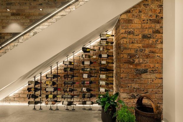 A compact wine display beneath a staircase, featuring rows of horizontally stored bottles on a black metal rack. Exposed brick walls, a white plastered stair underside, concrete flooring, linear recessed lighting, and potted greenery create an industrial cellar-like setting.