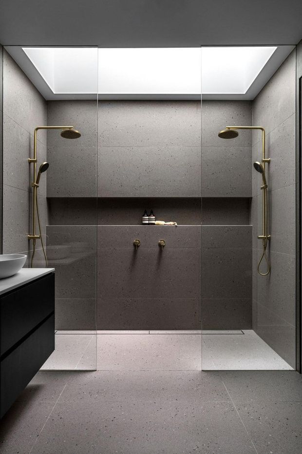 A contemporary bathroom with a symmetrical double walk-in shower enclosed by frameless glass. Large dark grey speckled stone-look tiles cover the walls and floor, with a recessed shelf, twin brushed-gold shower systems and controls, and a bright recessed ceiling light. A black floating vanity with a white basin is partially visible at left.