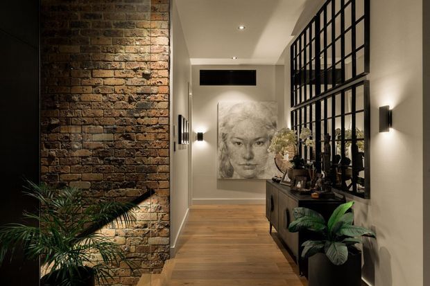 A refined interior hallway or entry passage featuring an exposed rustic brick feature wall, warm timber flooring, pale plaster walls, black-framed industrial-style glazed partitions, and a dark timber console with decorative objects and plants. Wall sconces and recessed downlights create a warm, softly illuminated atmosphere, while a large monochrome portrait artwork anchors the far end.
