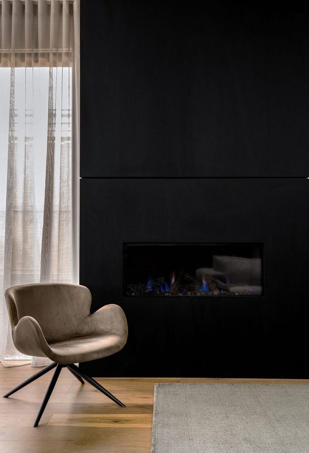A moody contemporary living room vignette with a black feature wall incorporating a long, built-in linear fireplace. A sculptural taupe lounge chair sits beside sheer full-height curtains, with pale timber flooring and a neutral textured rug.
