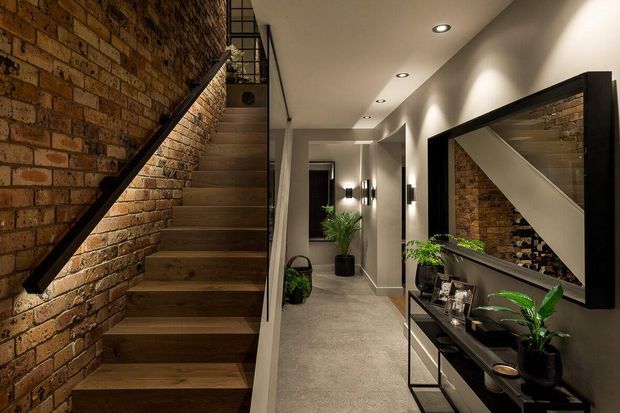 A refined residential hallway and stairway featuring a warm exposed brick stair wall, dark timber stair treads, black metal handrails, and a long corridor with recessed ceiling spotlights. A large black-framed mirror reflects the staircase, while a slim console with indoor plants and decorative objects runs along the opposite wall.