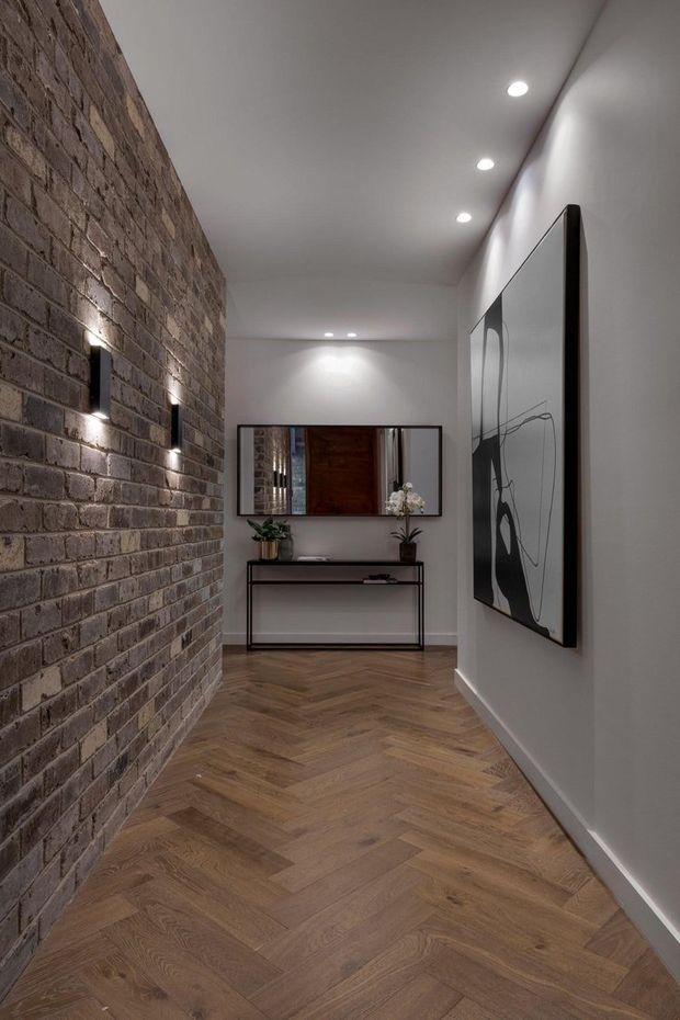 A narrow, contemporary hallway or entry passage with a warm herringbone timber floor, exposed brown brick feature wall with small wall lights, smooth white walls and recessed ceiling spotlights. A slim black console table, mirror, plant and abstract artwork create a refined entry vignette.