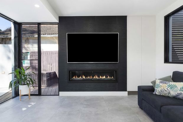 Bright contemporary living room with a large black feature wall incorporating a wall-mounted television and linear built-in gas fireplace. The room has pale large-format tiled flooring, white walls and ceiling, dark-framed glazing and doors opening to an outdoor deck, plus a dark sectional sofa and potted plant.
