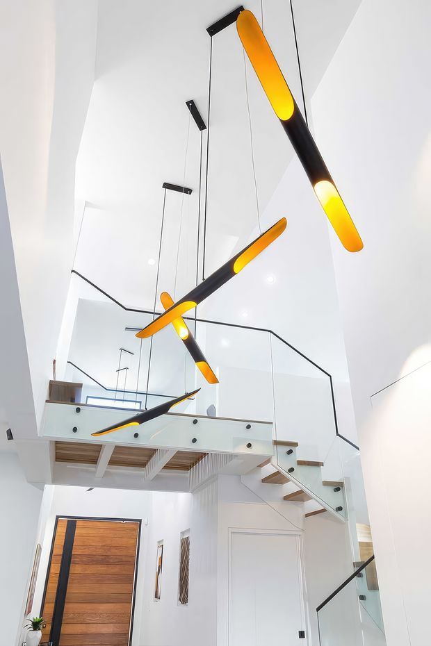 Bright contemporary double-height entry hall with a floating mezzanine and staircase. The white interior is defined by sculptural suspended pendant lights with black bodies and glowing amber ends, frameless glass balustrades with dark handrails, pale timber stair treads, and a large timber pivot front door. Skylight-like glazing and recessed ceiling lights create an airy, dramatic vertical space.