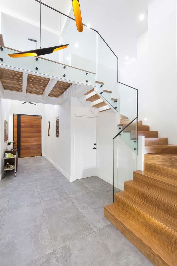 Bright contemporary residential entryway and stairway with a dramatic floating timber staircase, frameless glass balustrades with dark fittings, a mezzanine overhead, white walls, large grey floor tiles, and a timber front door. Sculptural orange and black pendant lights add a bold accent, while timber slatted ceiling detailing is visible beneath the upper level.