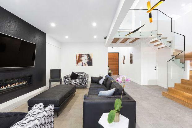 Bright contemporary open-plan living room with a black textured feature wall, wall-mounted television and linear fireplace, large dark sectional seating, and a sculptural coffee table. A floating timber staircase with frameless glass balustrades leads to a mezzanine, while white walls, recessed lighting, and pale flooring create a clean, spacious interior.