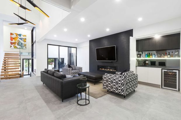 Bright contemporary open-plan living room with a high sloping ceiling, large pale tiled floor, black feature TV wall with linear fireplace, dark leather sectional seating, patterned armchair, built-in illuminated bar cabinetry and a nearby open-riser timber stair. Linear pendant lighting and recessed downlights reinforce the modern fit-out.