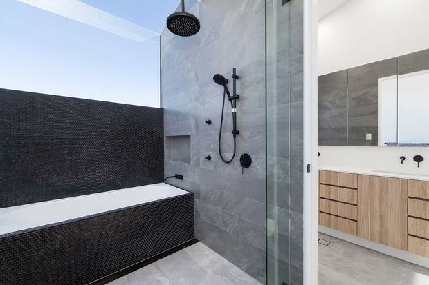 Contemporary bathroom featuring a freestanding-style bathtub clad in small dark mosaic tiles, a large grey stone-look tiled shower wall with rain shower and handheld fittings, and a clear glass shower screen. A timber-look floating vanity with white countertop and integrated basins is visible through the opening, along with a large wall mirror. The space has a clean, refined, neutral palette with strong black, grey, white, and light timber contrasts.