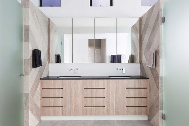 Contemporary bathroom featuring a wide double vanity with pale timber-look cabinetry, a dark stone benchtop, integrated basins and wall-mounted tapware. Large mirrored cabinetry spans the rear wall, while the side walls are finished in geometric timber-toned patterned tiles. Frosted glass doors frame the space.