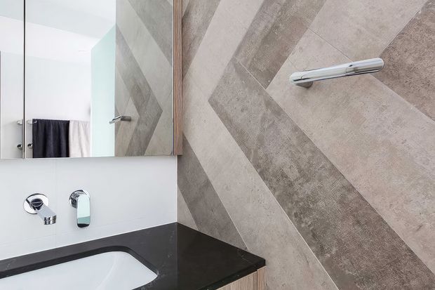 Close-up of a contemporary bathroom vanity and bathtub area. The space features a large mirrored wall cabinet, white wall surfaces, a black stone benchtop around the bathtub, chrome tapware, and a feature wall clad in diagonally arranged large-format stone-look tiles. A towel is visible in the mirror reflection.