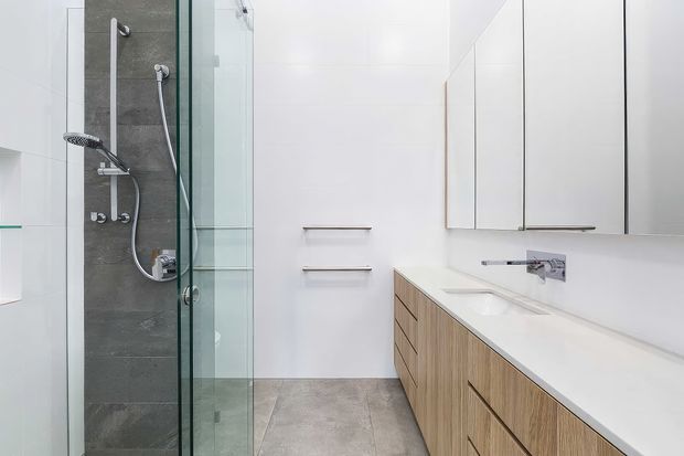 Bright contemporary bathroom with a frameless glass walk-in shower, dark grey tiled shower feature wall, wall-mounted shower fittings, a floating light-wood vanity with integrated white benchtop and basin, and white mirrored wall cabinetry. The room has a clean, minimalist appearance with large-format grey floor tiles and abundant white surfaces.