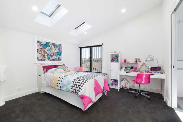 Bright, playful children's bedroom with a single bed dressed in colorful patterned bedding, a white desk and pink swivel chair, open shelving, wall art, dark carpet, and abundant natural light from a skylight and black-framed window.