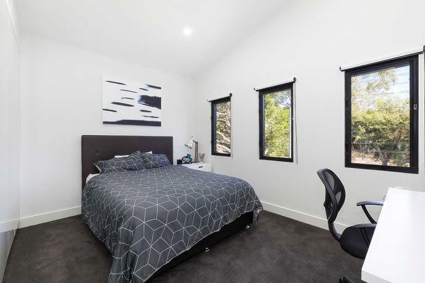 Bright, minimalist bedroom with a dark upholstered bed, charcoal geometric bedding, grey carpet, white walls and ceiling, black-framed windows with roller blinds, and a simple white desk with an ergonomic office chair. A monochrome abstract artwork hangs above the bed, while trees are visible through the windows.