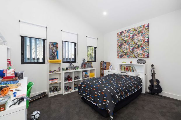 A bright, playful children's bedroom with three windows fitted with roller blinds, white walls, dark carpet, open shelving filled with toys and books, a double bed with a colourful patterned duvet, guitars, sports equipment, and a large multicoloured artwork above the bed.