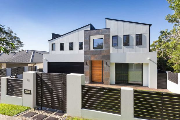 Street-facing view of a contemporary multi-storey residence with a geometric asymmetrical facade, white panel cladding, dark horizontal fencing, a charcoal garage door, timber front door, and a central stone-clad vertical feature. The home has varied rooflines, large rectangular windows, and a landscaped suburban frontage.