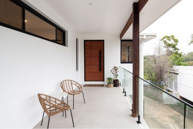 A bright, contemporary covered entry balcony or upper-level walkway leading to a timber front door. The space features white rendered walls, pale tiled flooring, dark timber structural posts and beams, a frameless glass balustrade, narrow black-framed clerestory glazing, and two woven timber lounge chairs. A neighbouring modern structure and landscaped surroundings are visible beyond the balustrade.