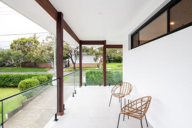 A bright covered outdoor terrace or balcony with a white tiled floor, frameless glass balustrade, dark timber structural posts and beams, and high horizontal black-framed windows. Two woven wicker lounge chairs overlook a landscaped suburban garden and lawn.